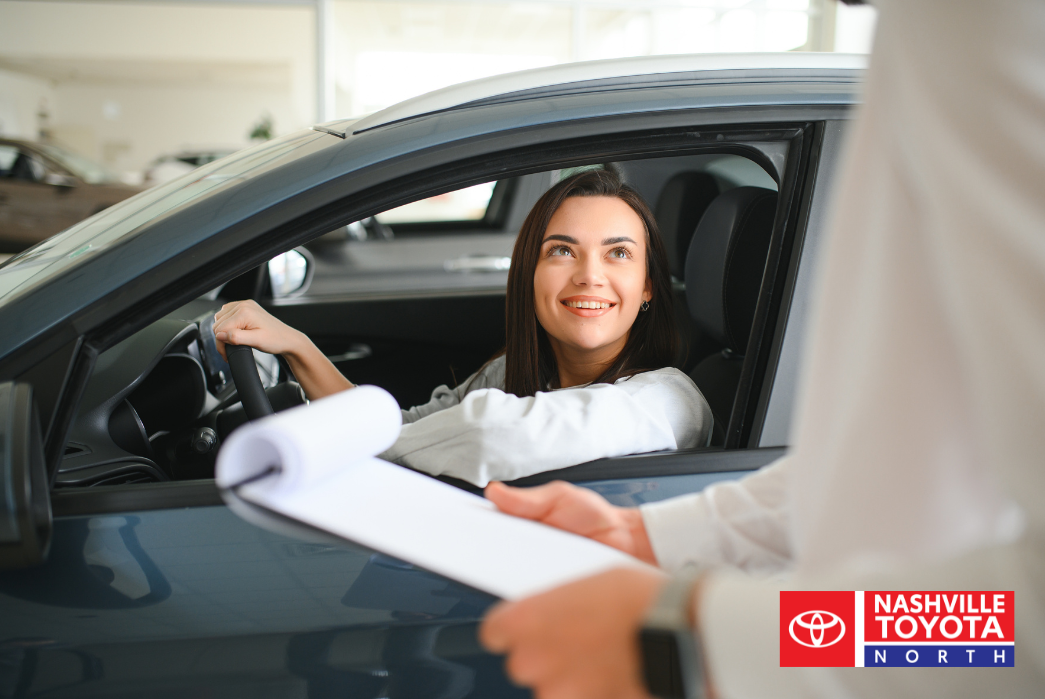 A person sits in the driver's seat of a car, looking up and smiling as someone with a clipboard stands outside the car.