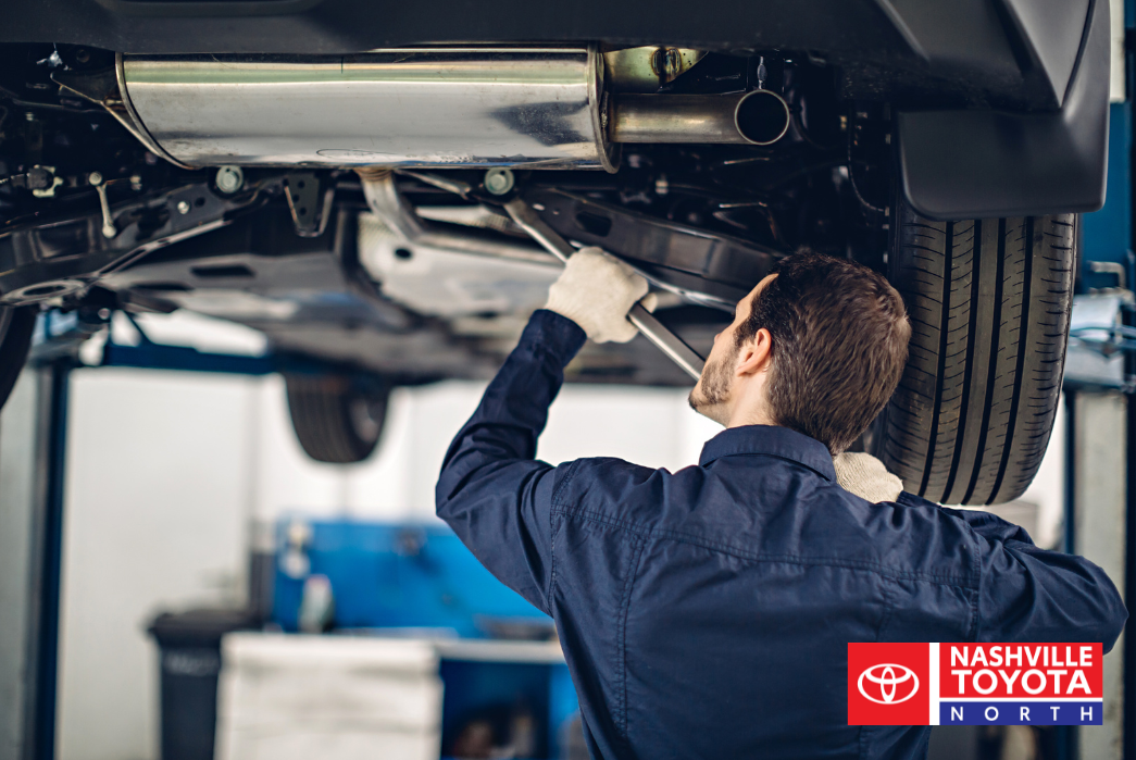 A mechanic wearing a blue uniform and white gloves works on the underside of a car that is raised on a lift, using a wrench.