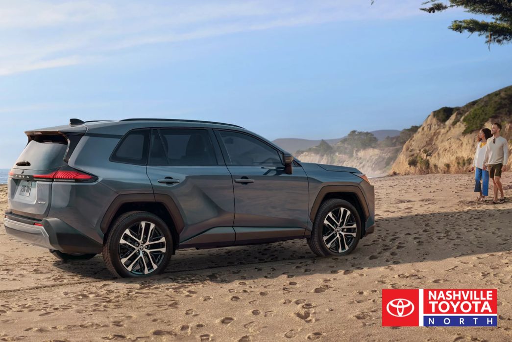 A dark blue SUV is parked on a sandy beach next to the ocean, with two people standing on the sand looking out at the water and a cliffside in the background.