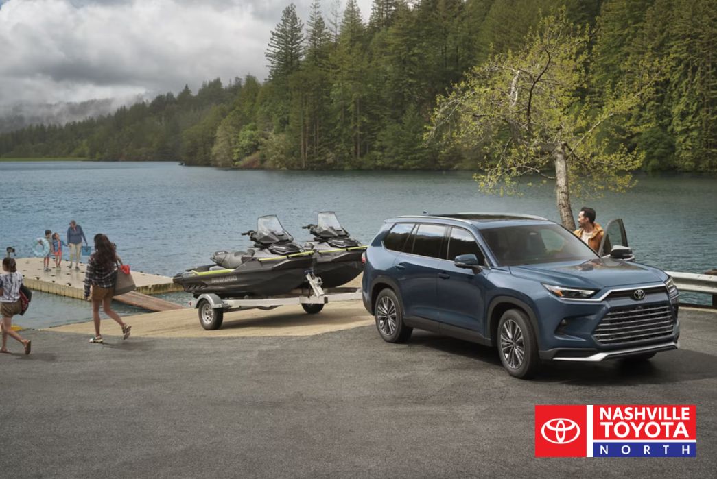 A dark blue SUV is parked next to a trailer carrying two jet skis by a lake with a forested background. People are walking on a dock towards the water.