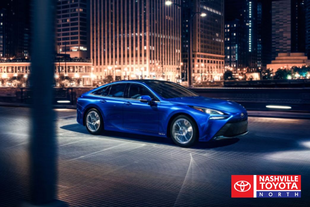 A blue sedan is parked on a street at night with city buildings illuminated in the background.