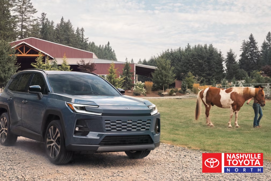 A blue SUV is parked on a gravel road in front of a farm with a red barn, while a person leads a brown and white horse in a grassy field.